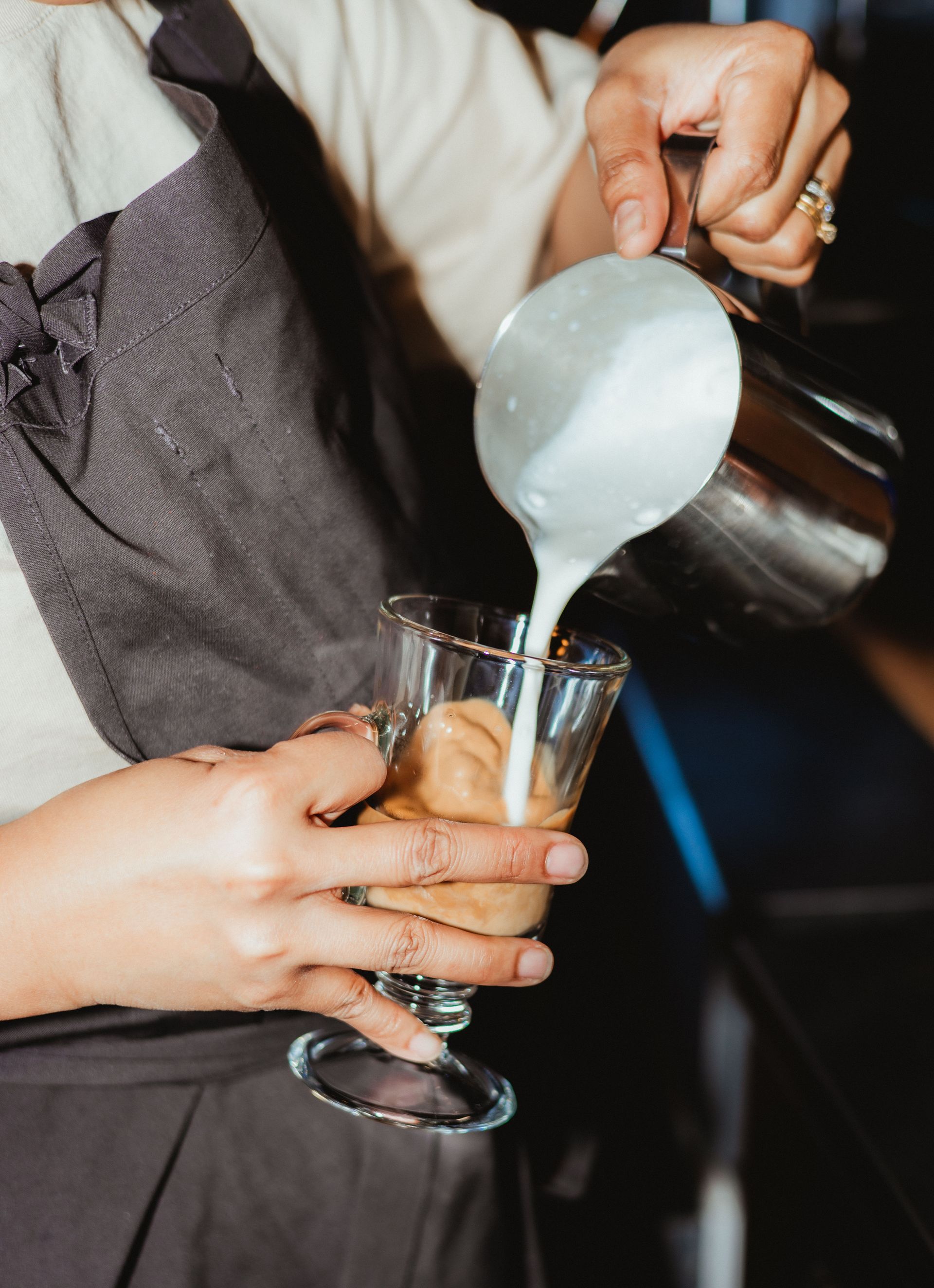 Henkilö kaatamassa maitoa hopeisesta kannusta kirkkaaseen lasiin ja valmistamassa juomaa.