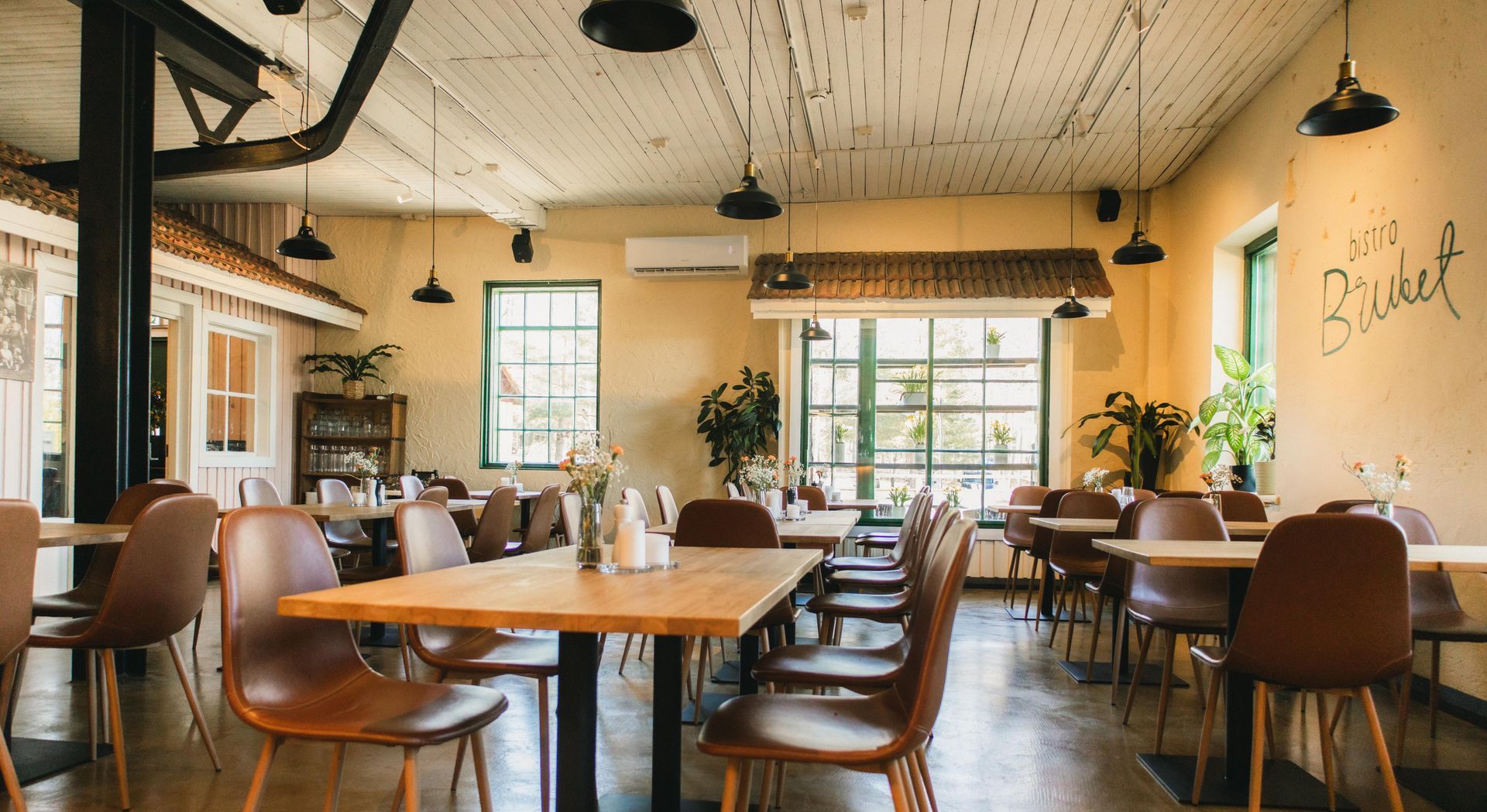 Ravintolan sisustus, jossa on puisia pöytiä ja tuoleja, luonnonvaloa ja vihreitä kasveja.