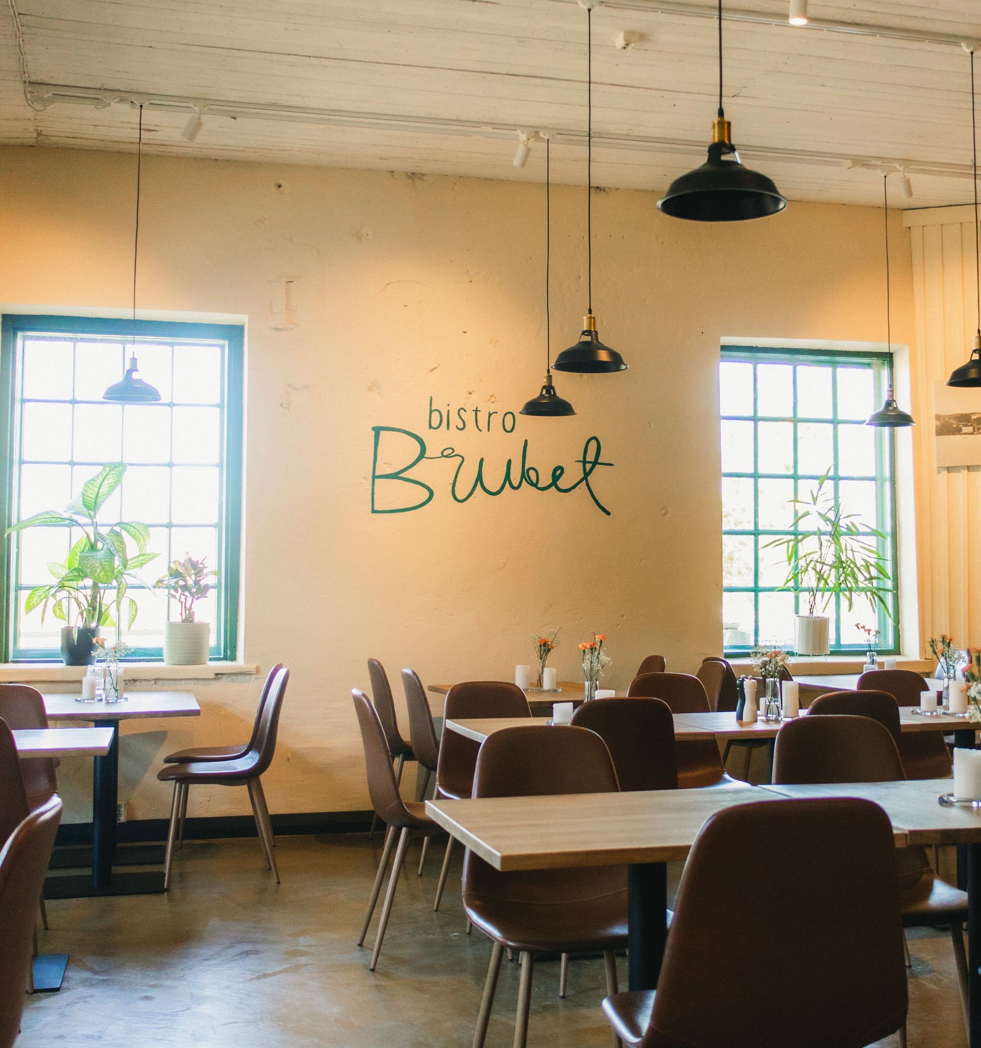 Ravintolan sisustus pöytineen, tuoleineen, ikkunoineen ja riippuvalaisimineen. Seinällä lukee