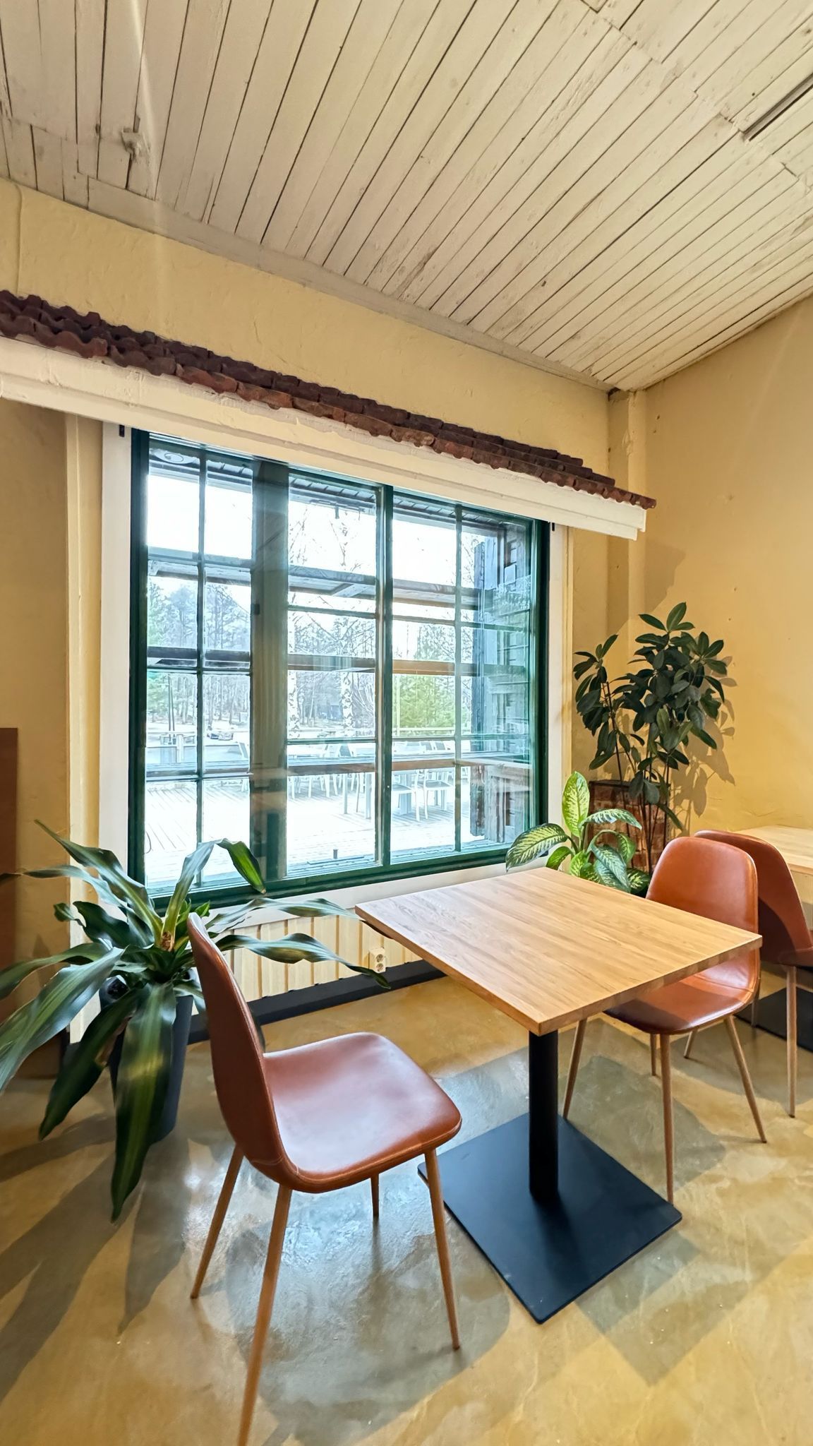 Kahvilan sisustus, jossa on neliönmuotoinen puinen pöytä, kaksi ruskeaa tuolia ja suuri ikkuna.