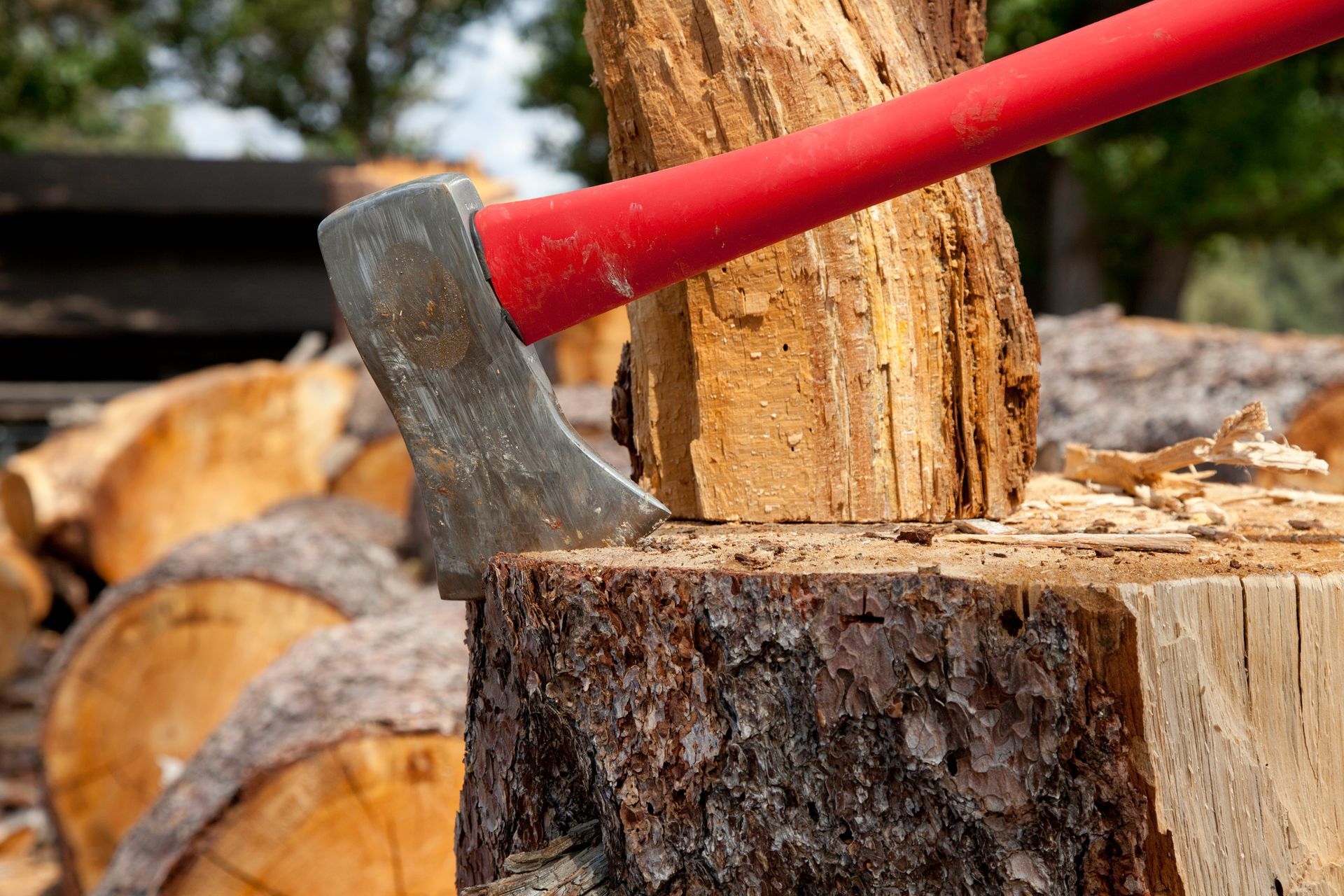 Red ax embedded in a wooden stump, ready to split wood; wood pile in background.