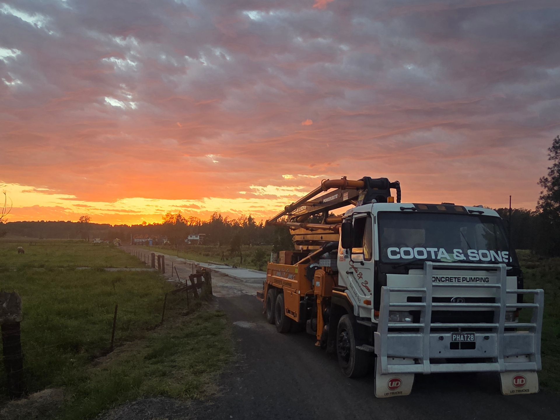 Concrete pump truck on a dirt road at sunset. Orange and white truck, dramatic sky over a rural scene.