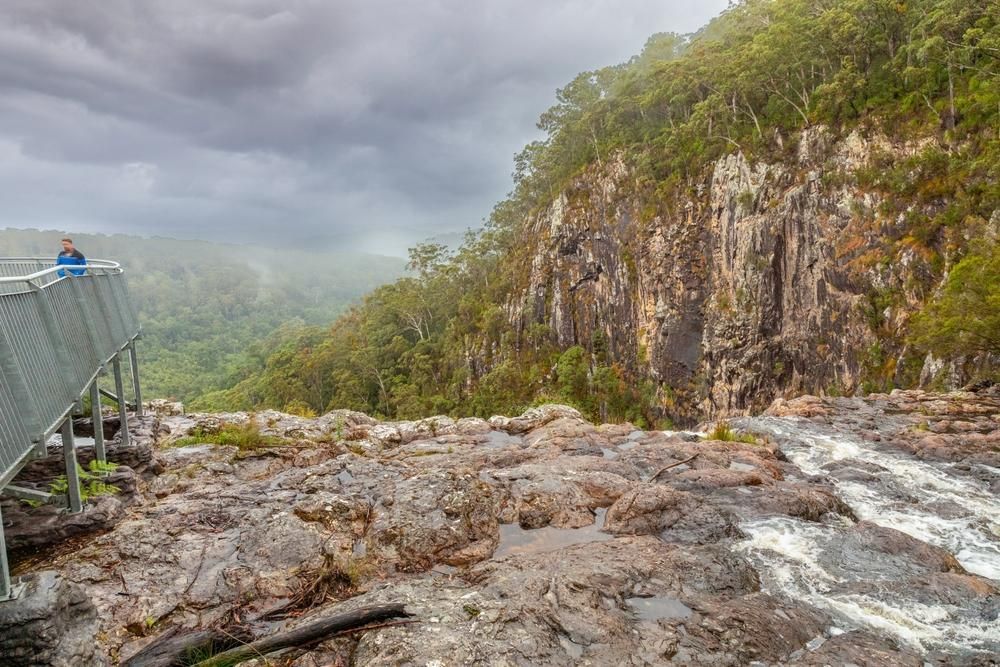 A Man Is Standing On A Bridge Overlooking A River — JBS Mobile In Alstonville, NSW