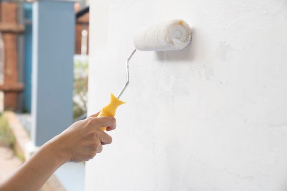 Hand using a paint roller on a white wall. Yellow handle, exterior setting.