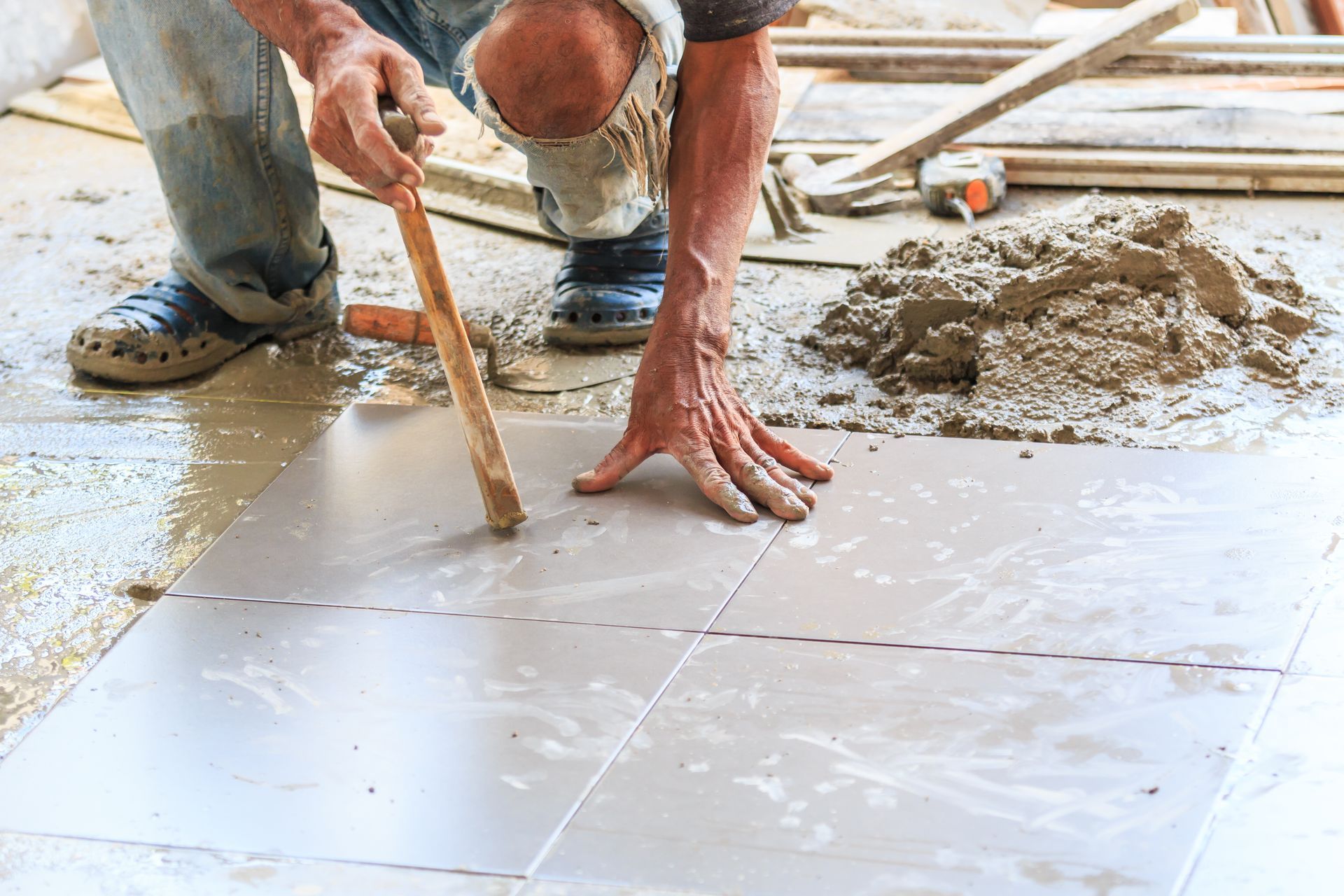 Man laying floor tiles, using a tool. Cement and building materials are present.