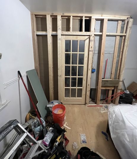 Interior room under renovation; wooden frame, French door, tools, and debris clutter floor.