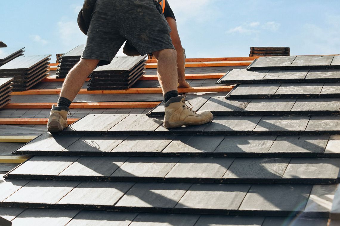 Roofer installing dark gray tiles on a roof, sunny day, wearing shorts and work boots.