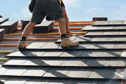 Roofer installing dark gray tiles on a roof, sunny day, wearing shorts and work boots.