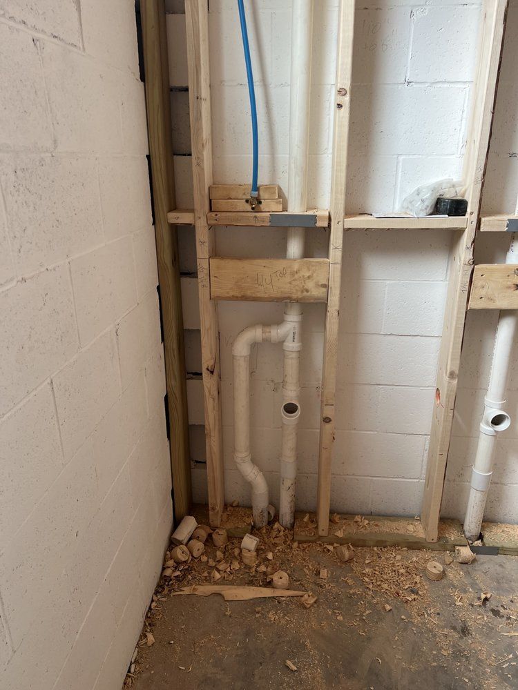 Framing and plumbing pipes inside a wall, near a concrete block wall, with wood shavings on the floor.