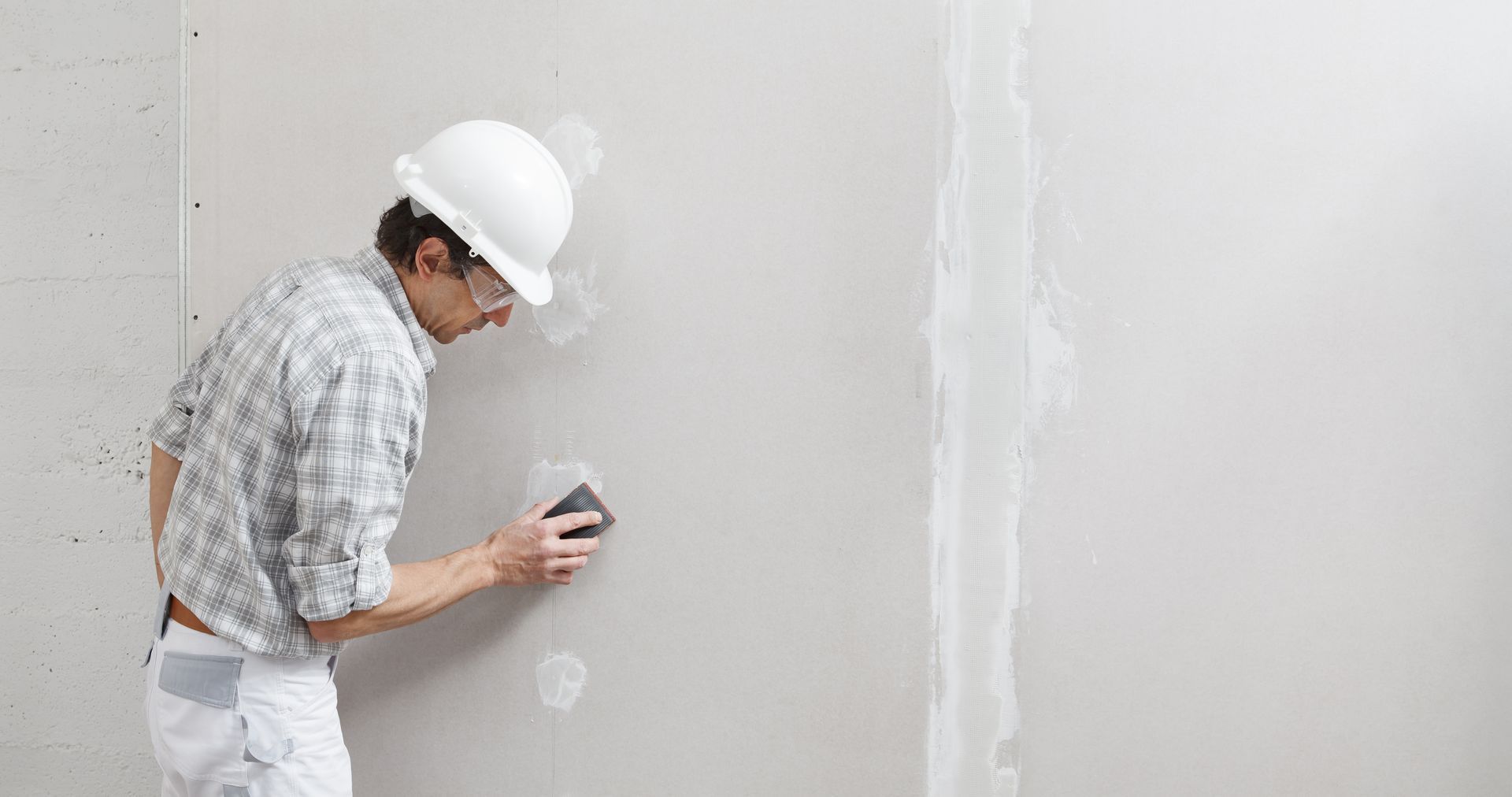 Man in hard hat sanding a patched section of drywall on a white wall.