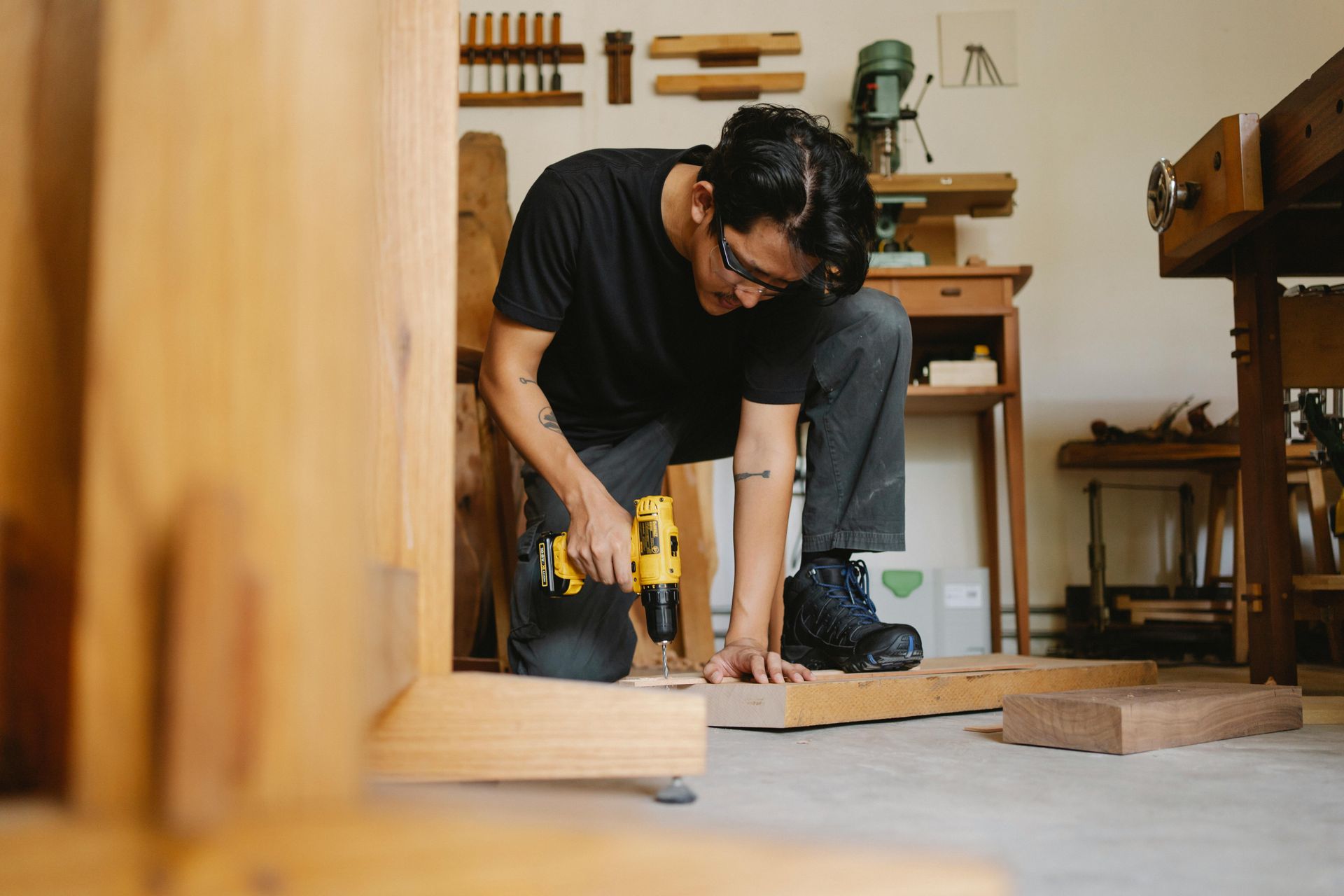 A person in safety glasses kneels in a woodshop, using a yellow power drill on a piece of wooden lumber.