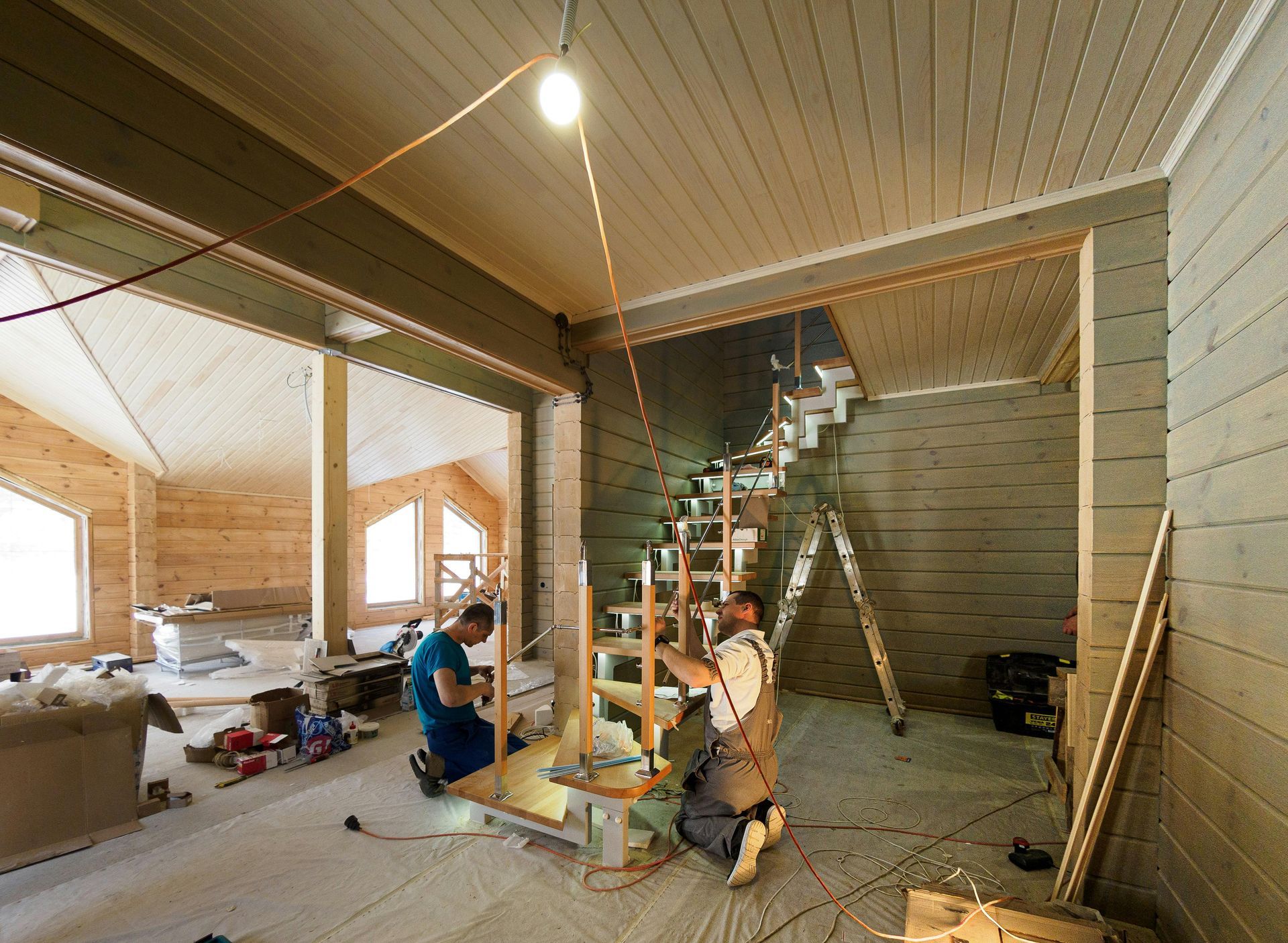 Interior under renovation with exposed staircase, wood-paneled walls, and workers installing a floor beam