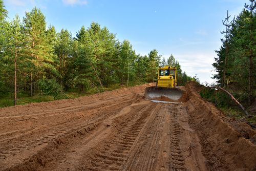 Bulldozer clearing a path in a forest, creating a dirt road. Yellow machine on brown earth, surrounded by green trees.