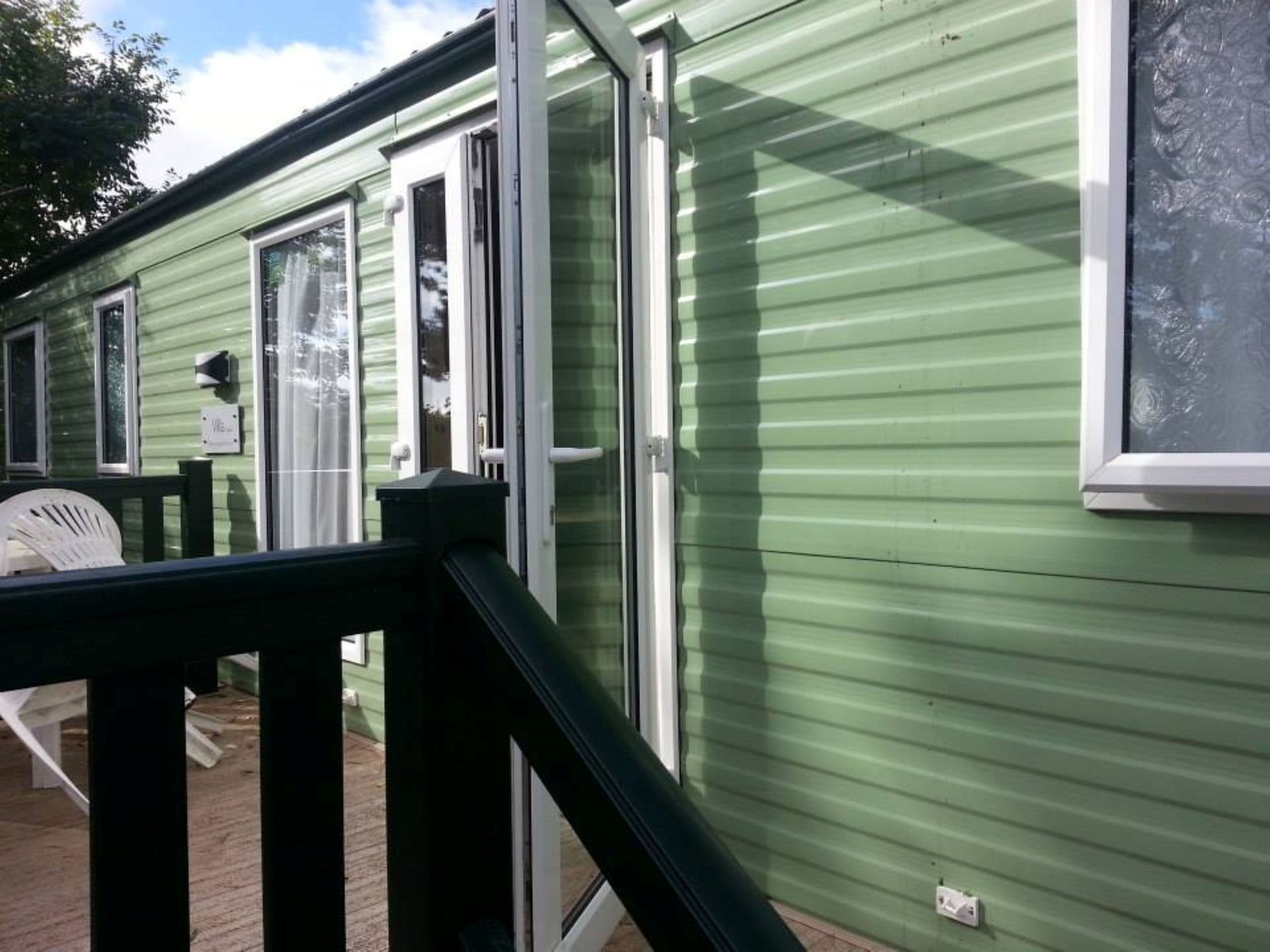 A side view of a light green mobile home with a dark railing on a deck and open glass patio doors.