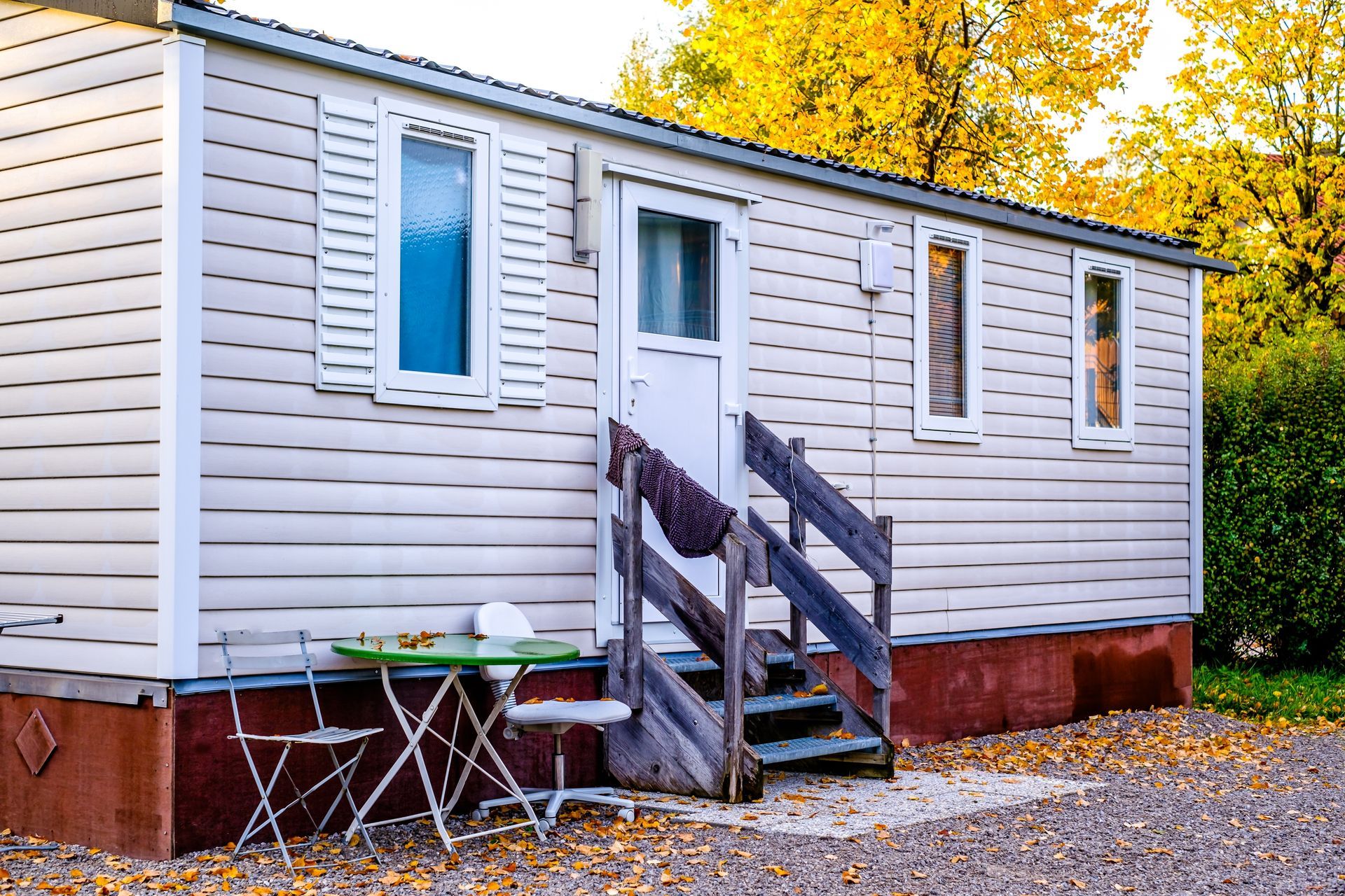 A light-colored mobile home with steps, a small green bistro table, and two chairs on a gravel patch near autumn trees.