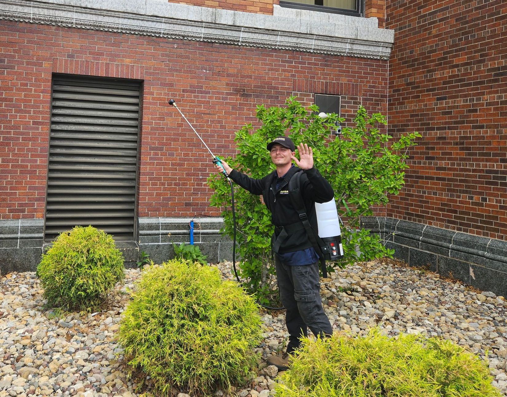 Person waving in a brick courtyard with shrubs and a dark vented door.