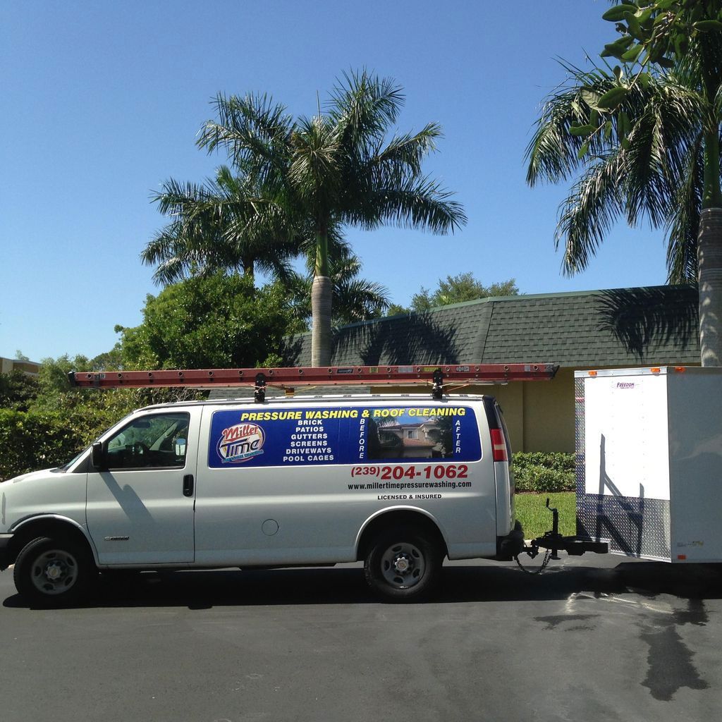 White van with a trailer parked on a driveway, displaying a business logo and phone number, palm trees and a building are in the background.