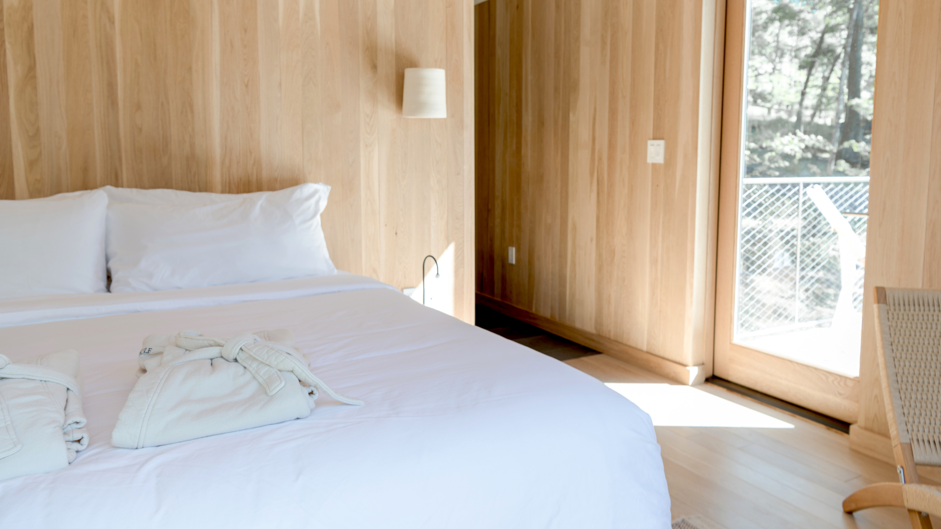 A minimalist bedroom featuring light wood paneled walls, a bed with white linens, and a bright window overlooking trees.