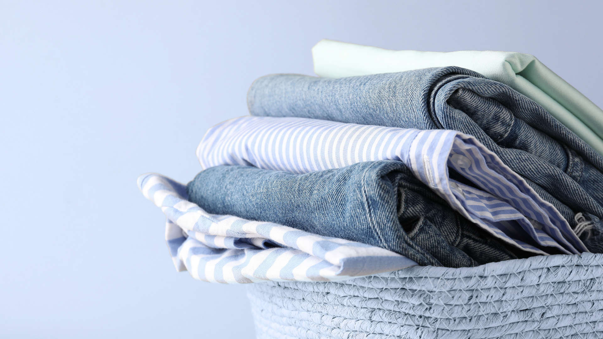 A stack of folded blue jeans and light-colored striped shirts rests in a textured basket against a pale blue background.