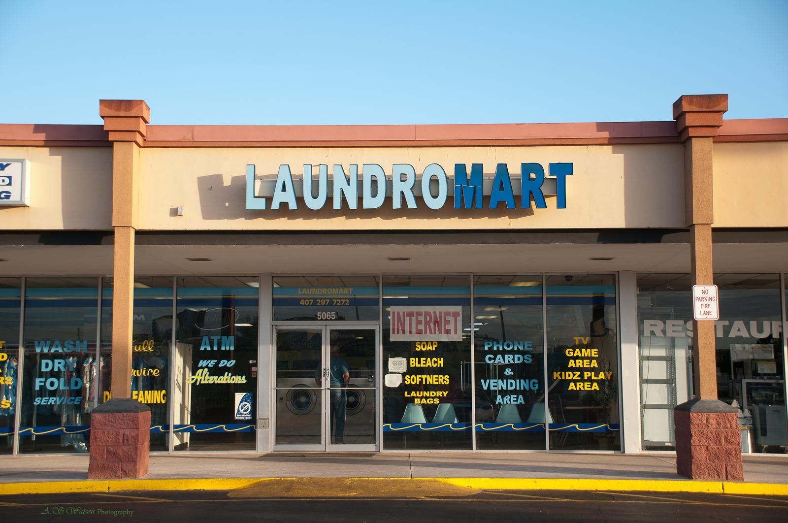 A beige strip mall storefront for Laundromart with large glass windows and blue signage under a clear blue sky.