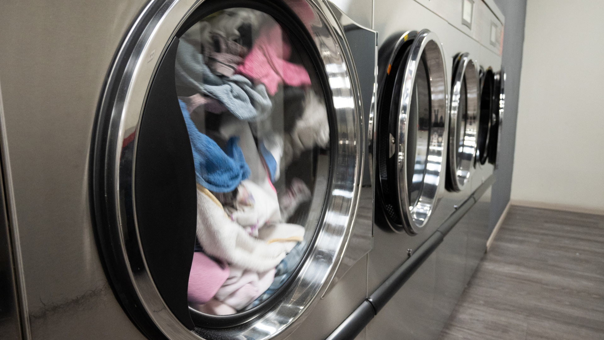 A row of stainless steel commercial dryers in a laundromat, with clothes tumbling inside the nearest unit.