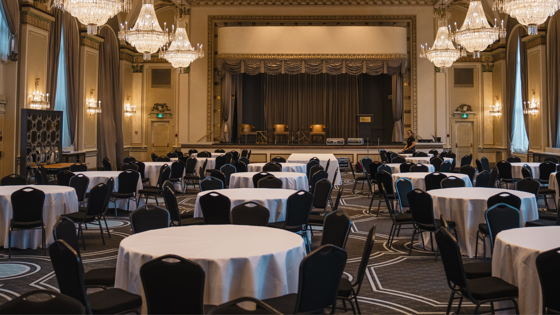 A grand ballroom set with round white tables and black chairs, facing a raised stage under crystal chandeliers.