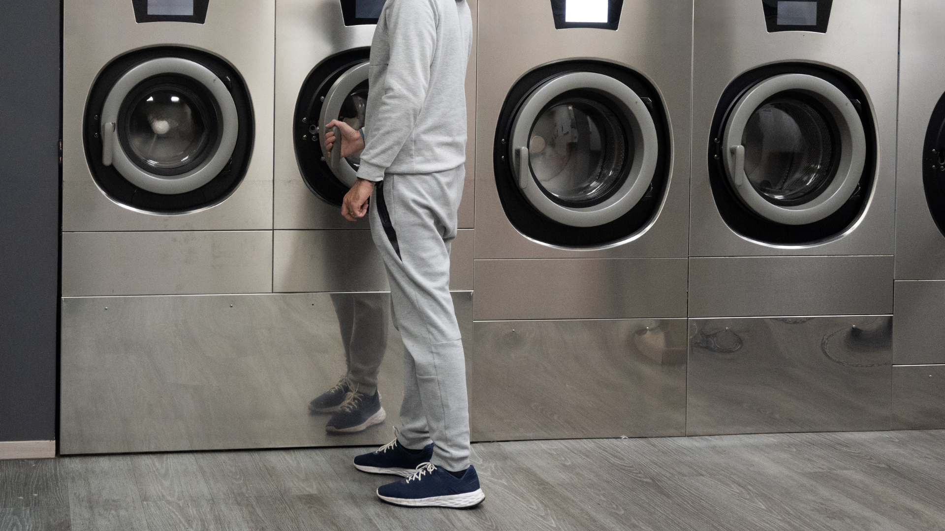 A person in a grey tracksuit stands in a laundromat, opening the door of a stainless steel washing machine.