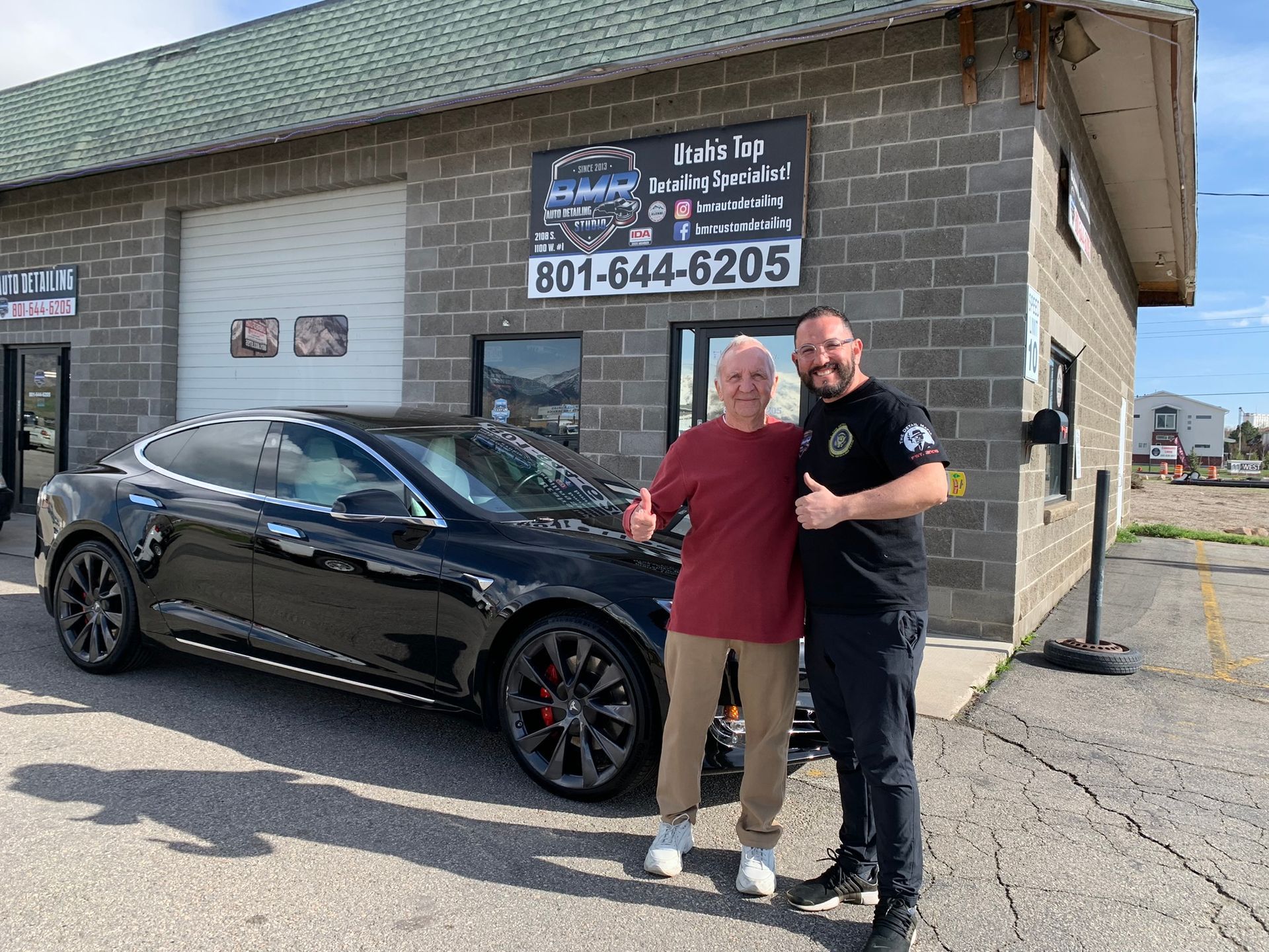 Man and customer stand in front of an auto shop with a black Tesla. Both give thumbs up.