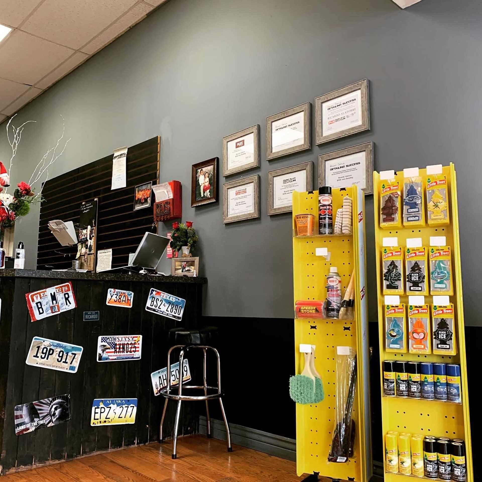A car detailing shop interior, with a black counter, yellow shelves, and framed documents on a gray wall.