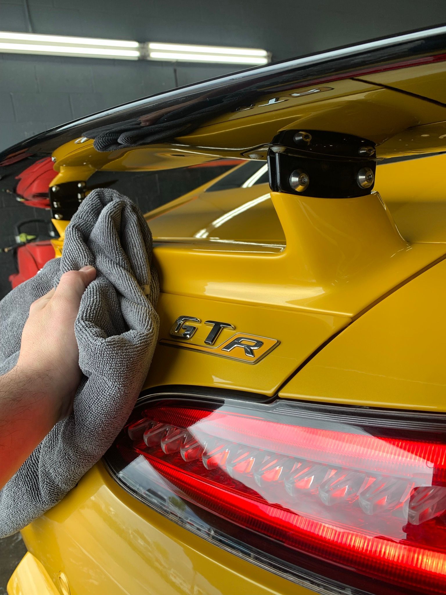 Hand wiping a bright yellow sports car's spoiler with a gray cloth; the word 