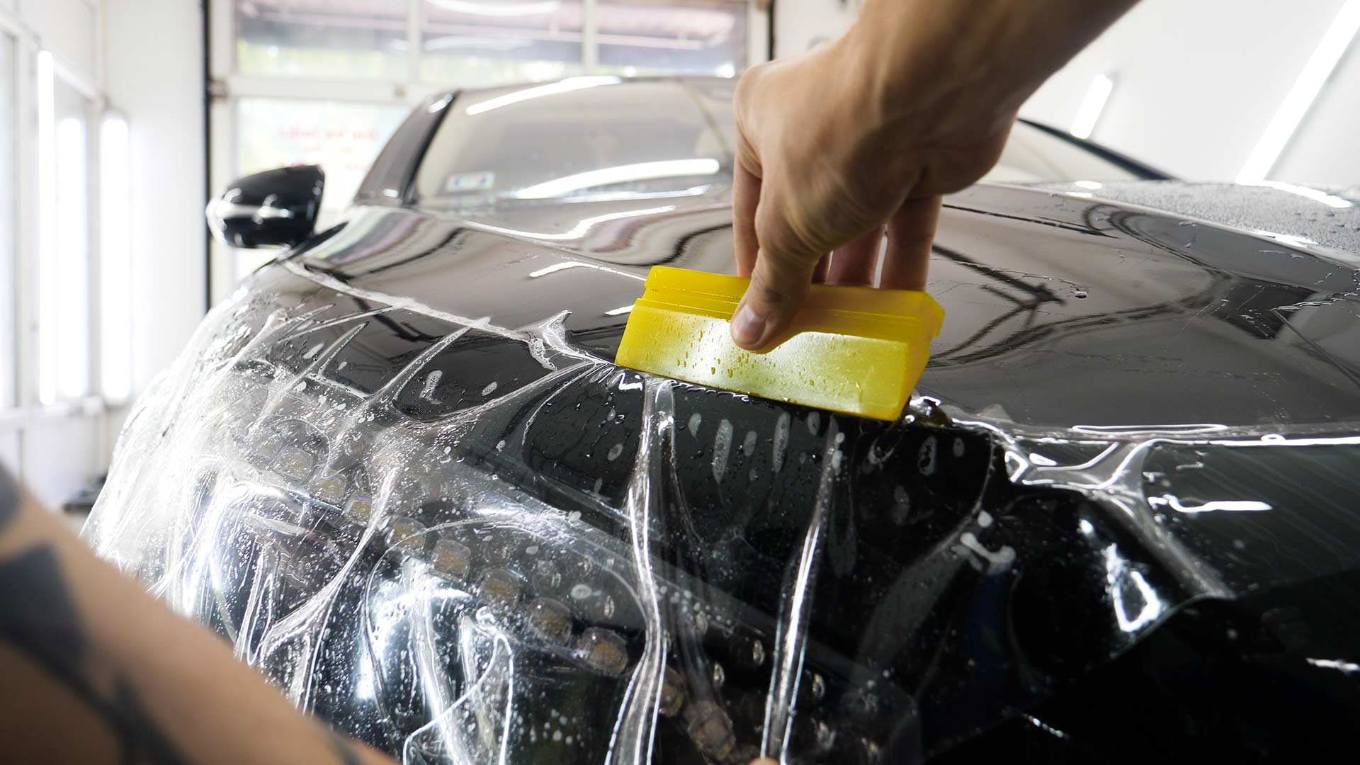 Person using a yellow squeegee to smooth protective film on a black car's front hood, inside a shop.