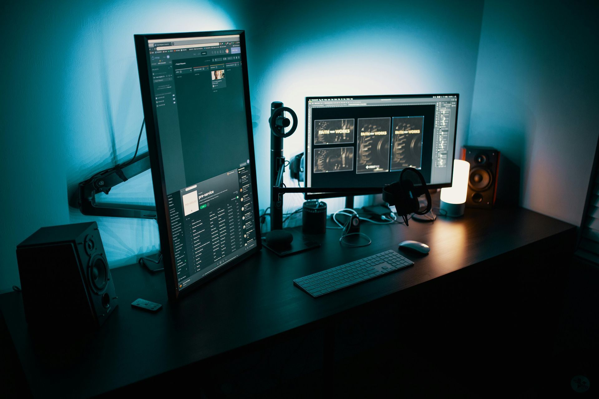 Dark workspace with two monitors, keyboard, speakers, and lamp. The monitors are displaying a dark user interface.