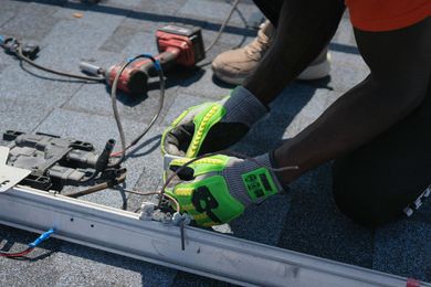 Worker in neon gloves using a power tool to install roof shingles on a rooftop.