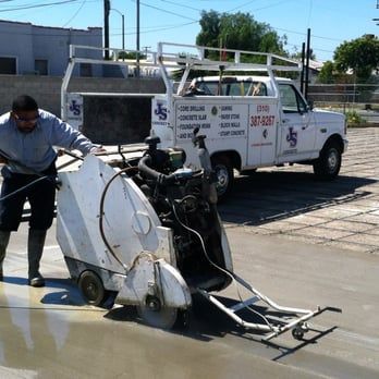 A construction worker uses an angle grinder to cut through a concrete surface, creating a large cloud of dust.