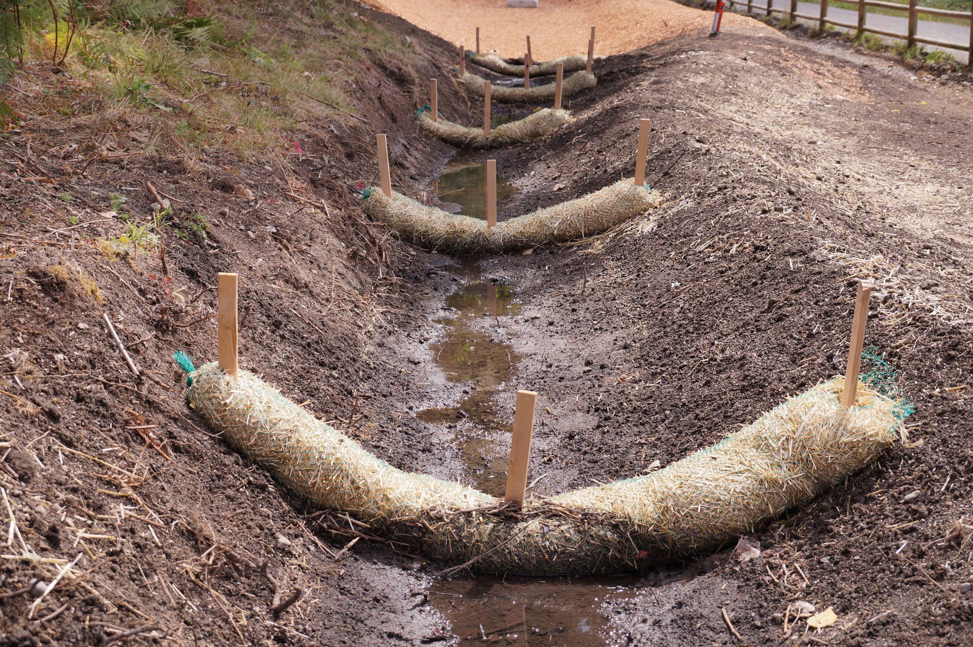 Two people work on a stone retaining wall in a yard, installing a vertical support pipe amid concrete blocks and gravel.