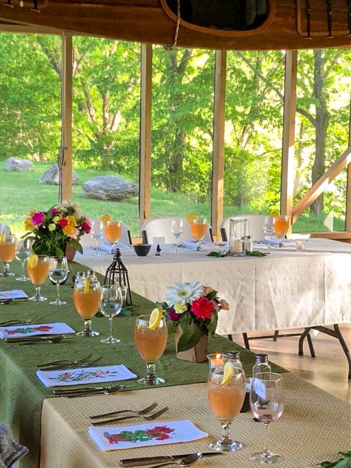 Dining table set for a meal, with floral centerpieces, drinks, and a green outdoor view through large windows.