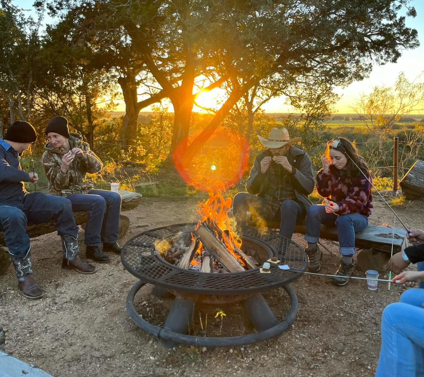 People around a fire pit roasting marshmallows at sunset.