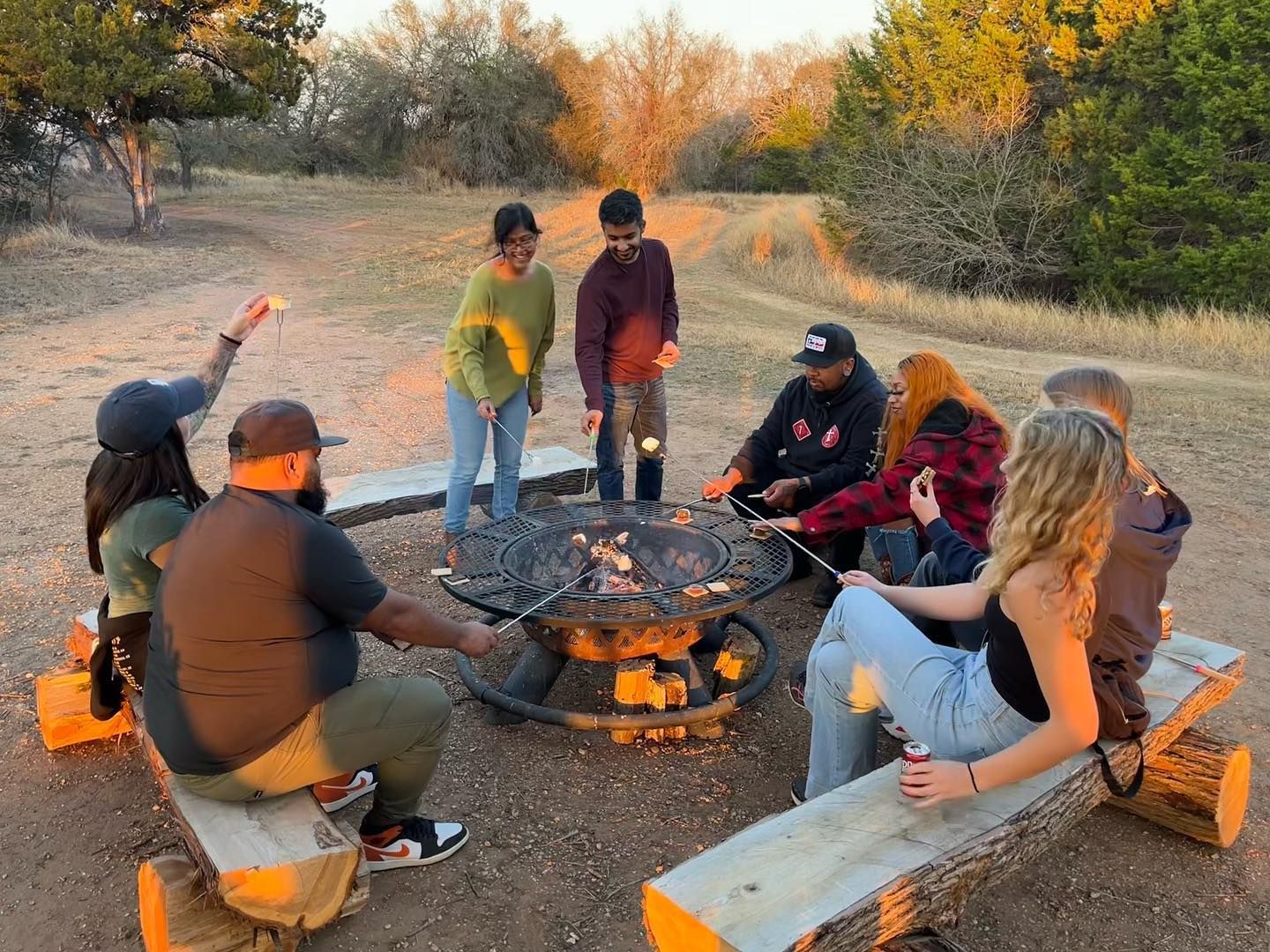 People roast marshmallows around a fire pit outdoors on wooden benches at sunset.