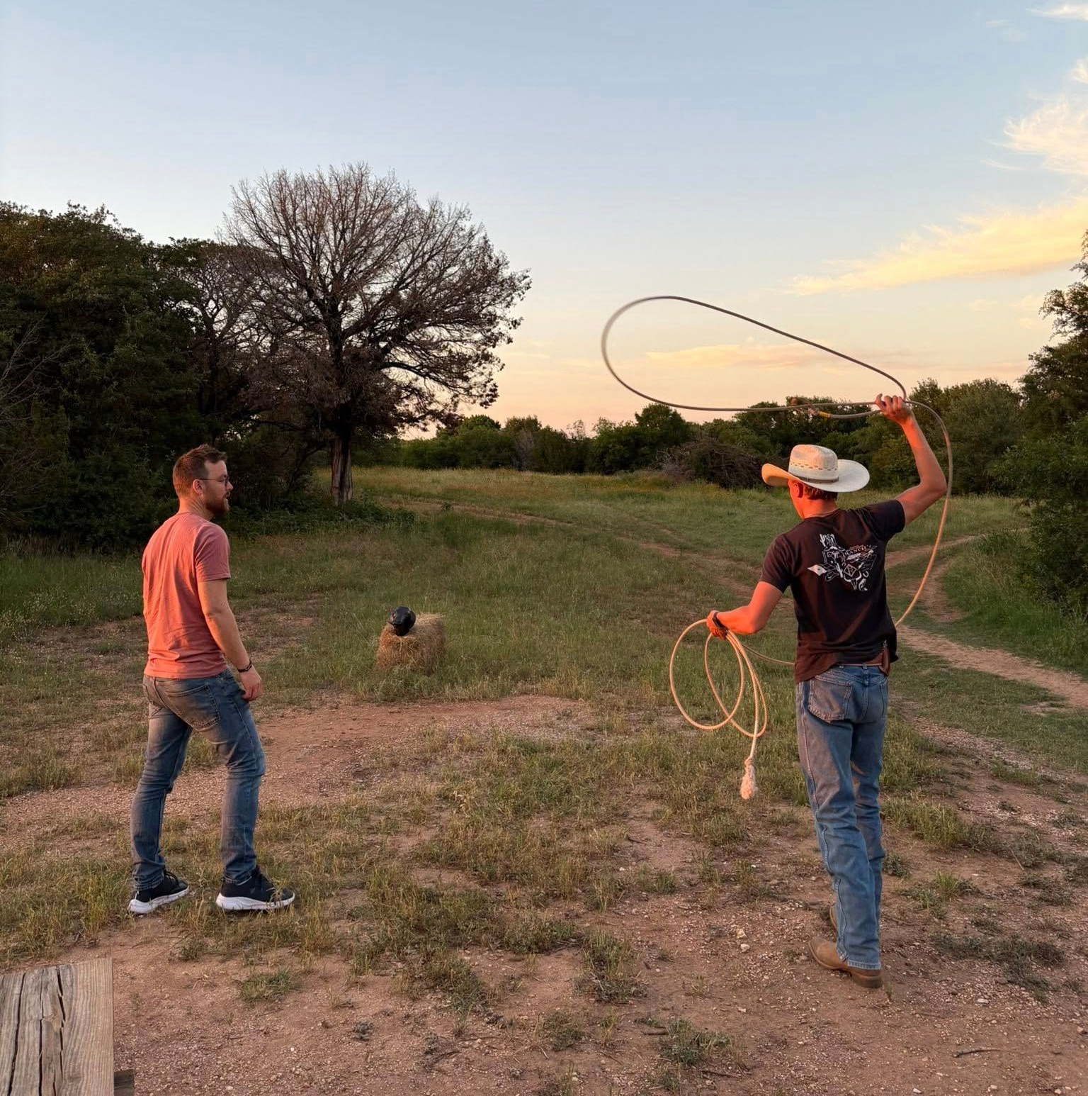 Man with a lasso, preparing to rope a target while another man watches on a grassy field at dusk.