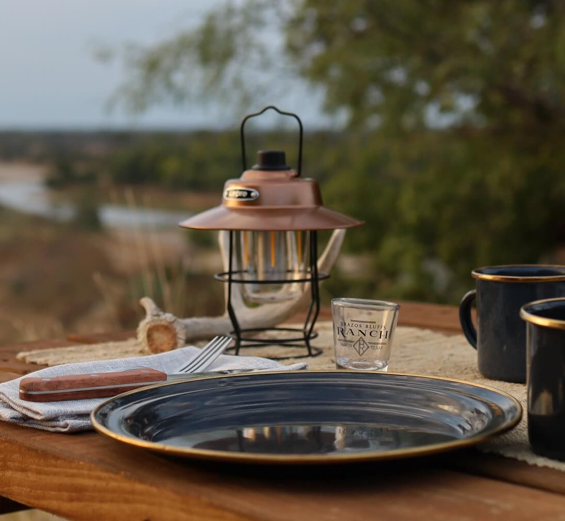 Camping table setting: lantern, plate, mugs, shot glass with river view.