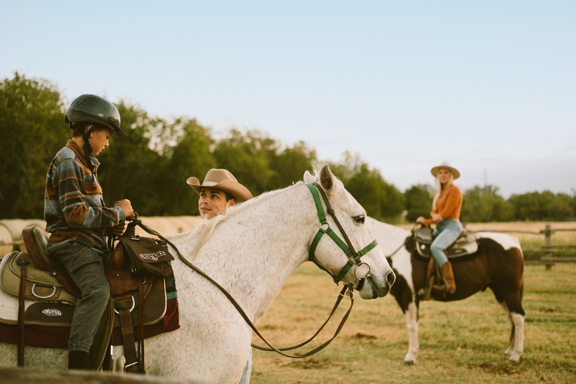 Man in cowboy hat leans against a rustic fence in front of a barn.