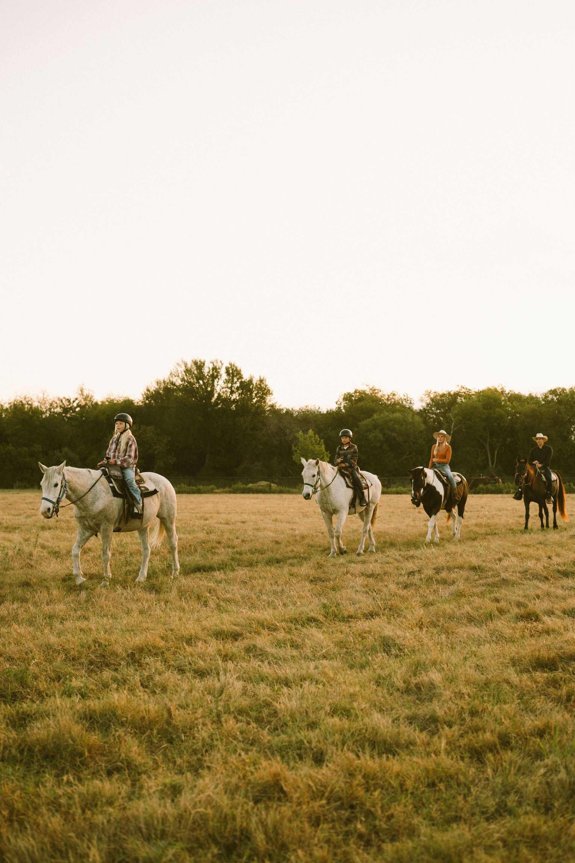 People horseback riding in a field, with trees in the background under an overcast sky.