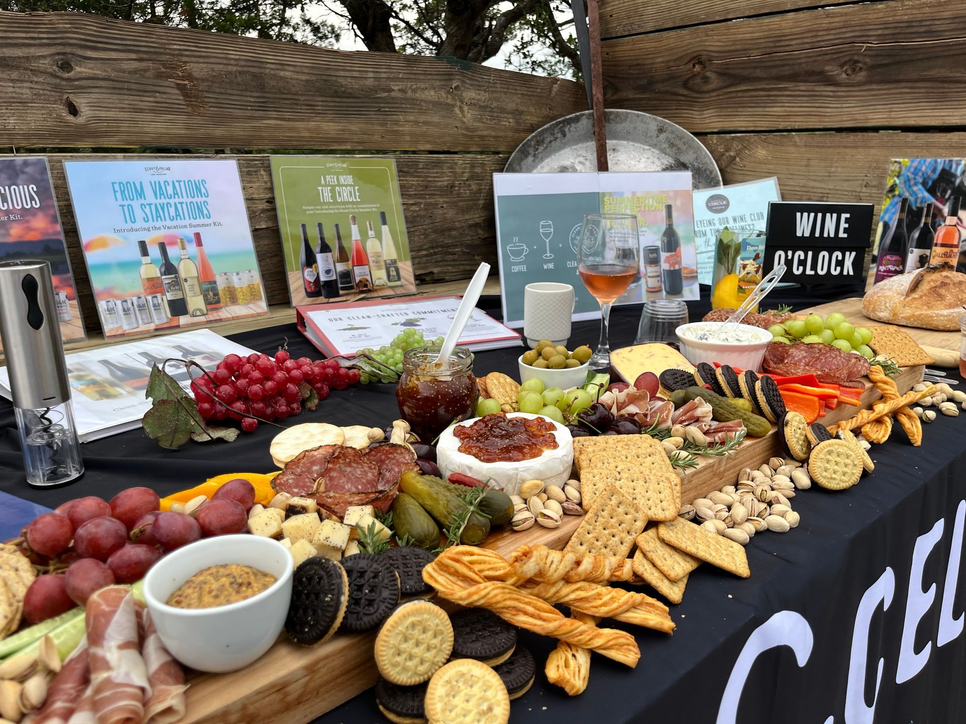 Table of food and books, with wine, cheese, crackers, fruit, and snacks, set outdoors.