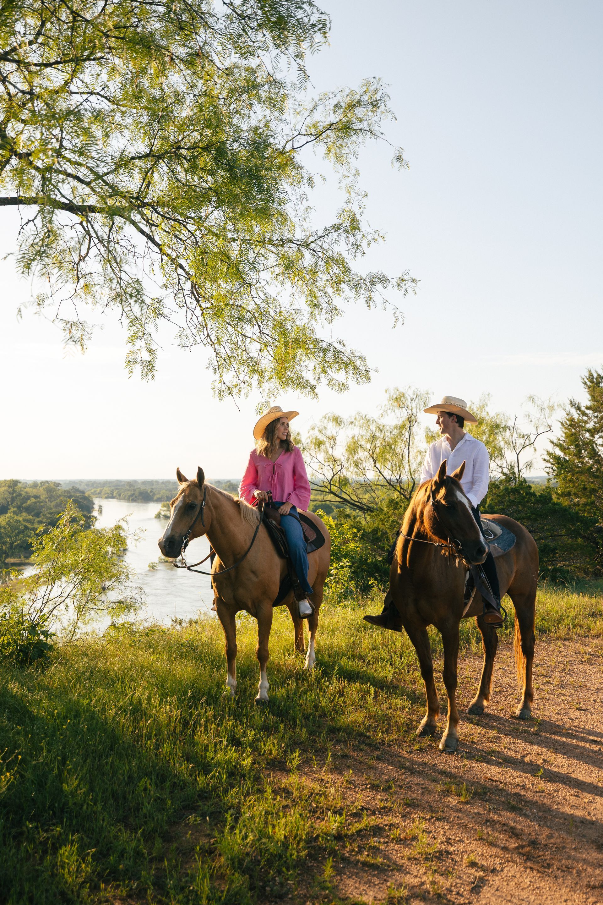 Two people on horseback, wearing hats, overlooking a river. Sunlight bathes the scene.