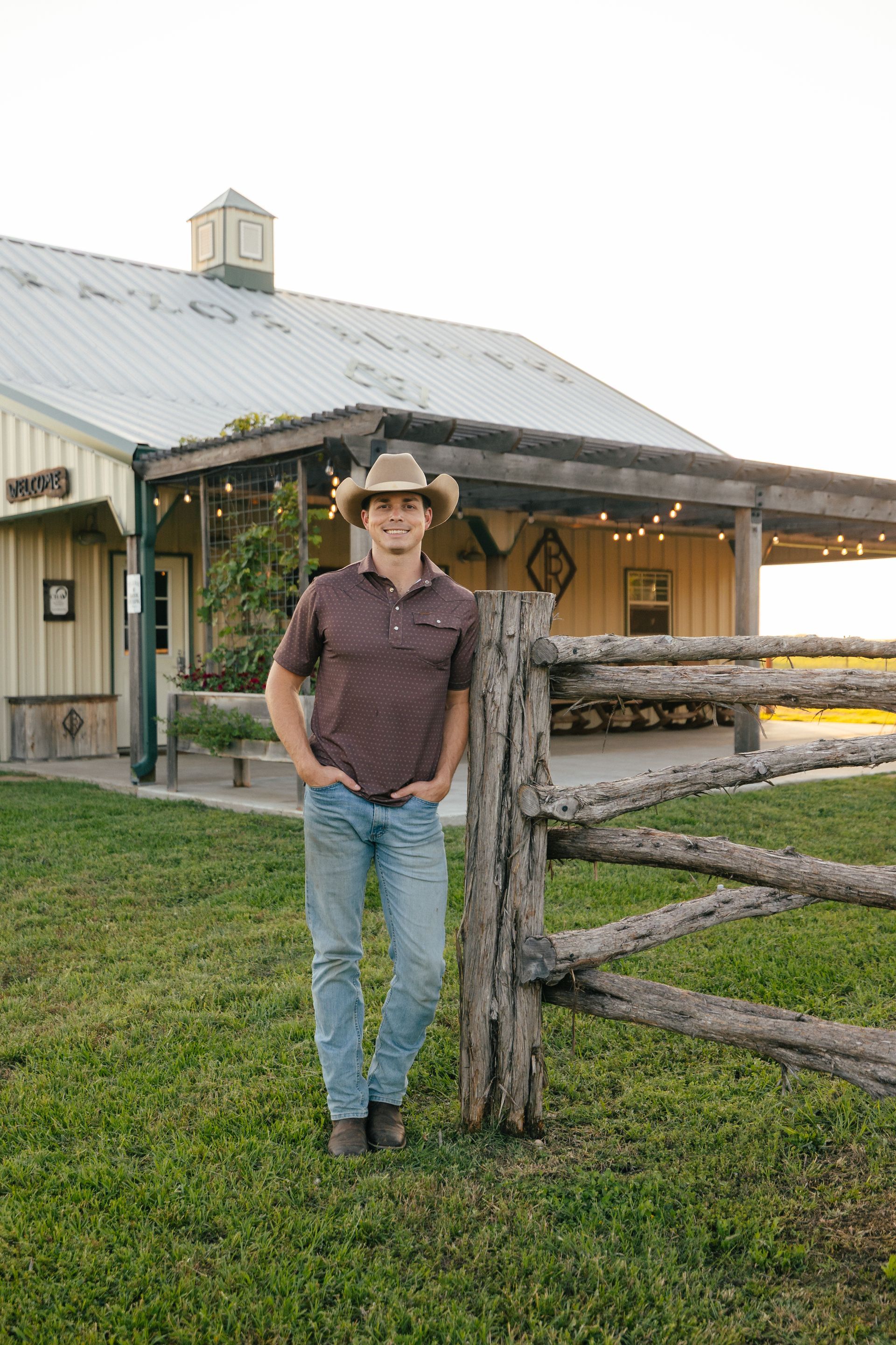 Man in cowboy hat leans against a rustic fence in front of a barn.
