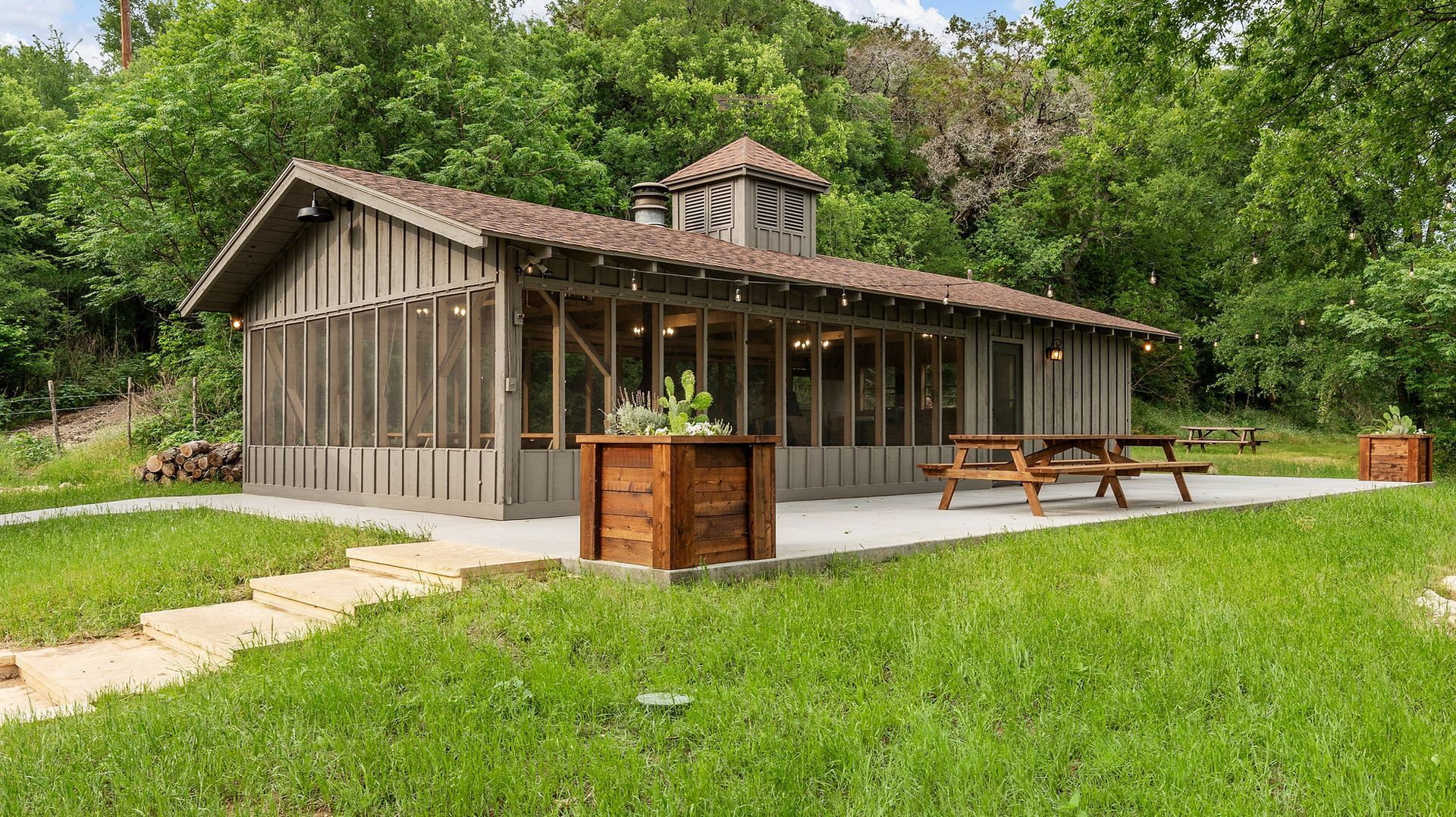 Screened-in wooden structure with picnic tables on a concrete patio, surrounded by green grass and trees.