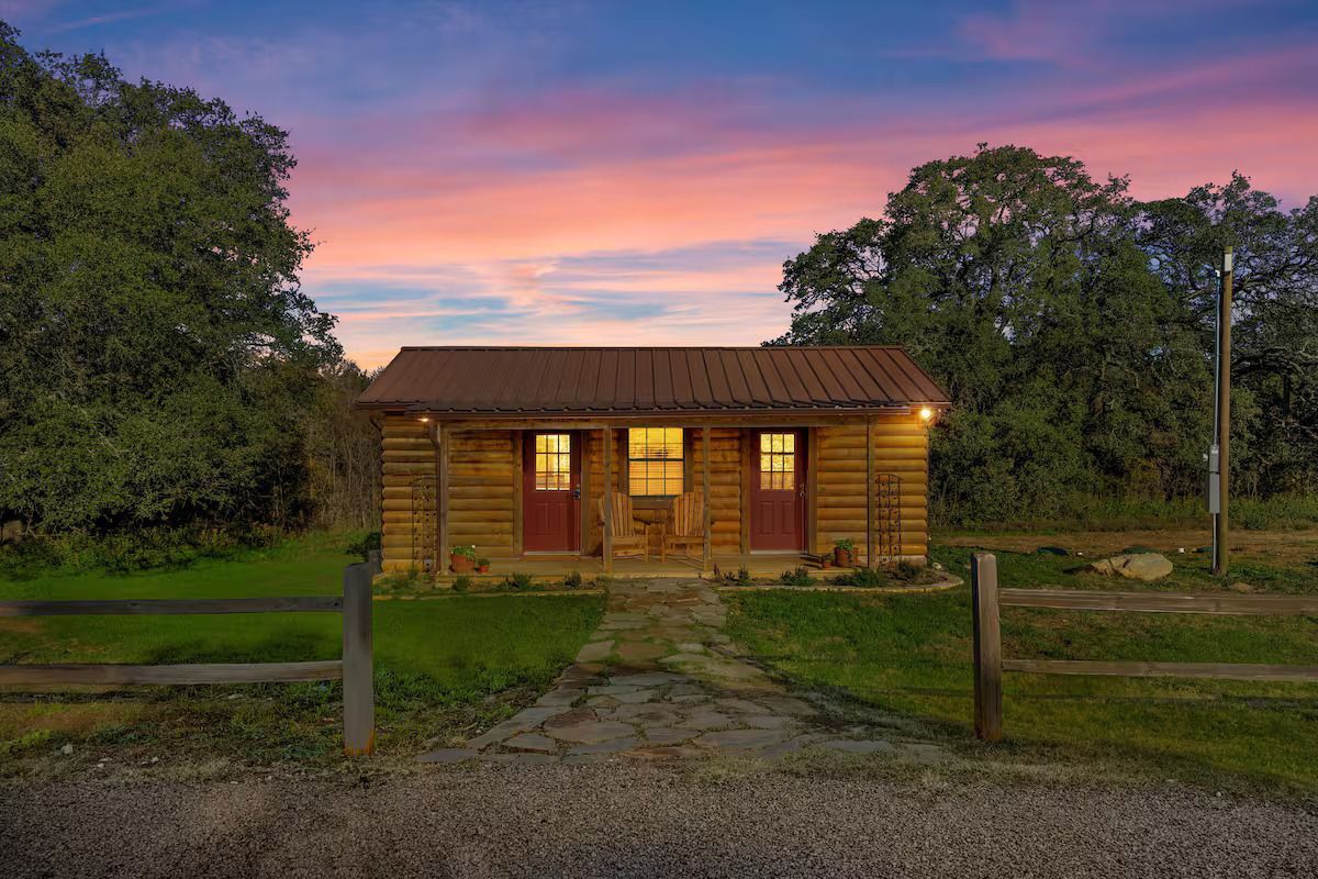 Wooden cabin with red doors at dusk, framed by trees and a colorful sky.