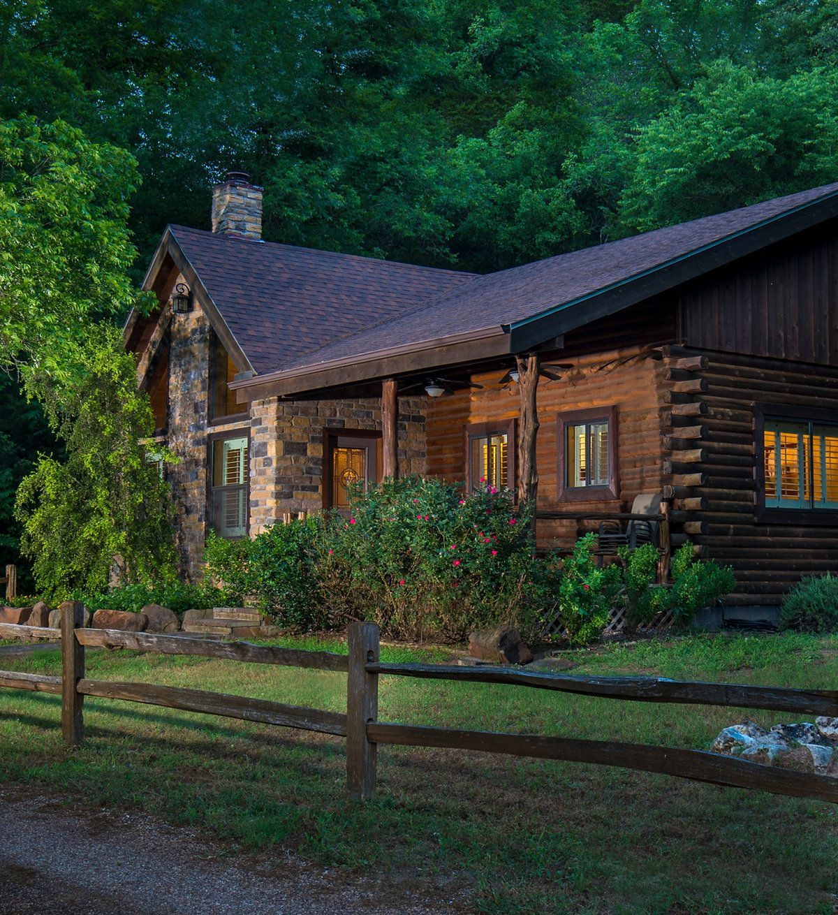 Rustic log cabin with stone chimney, porch, and wooden fence surrounded by green foliage.
