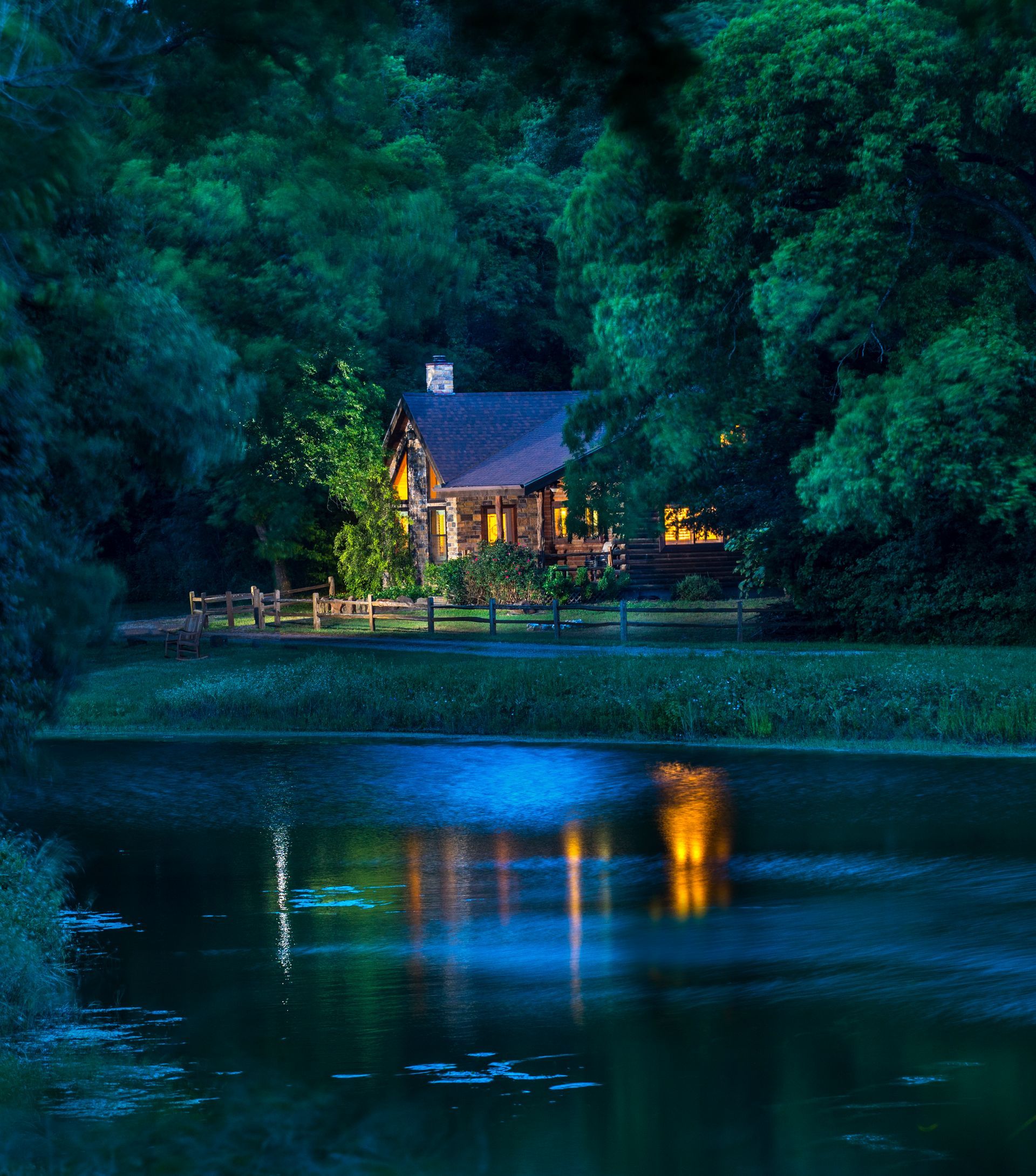 Cozy stone cabin glowing at dusk beside a calm pond, surrounded by dense trees and reflecting warm lights across the water at Brazos Bluffs Ranch in Waco, Texas.