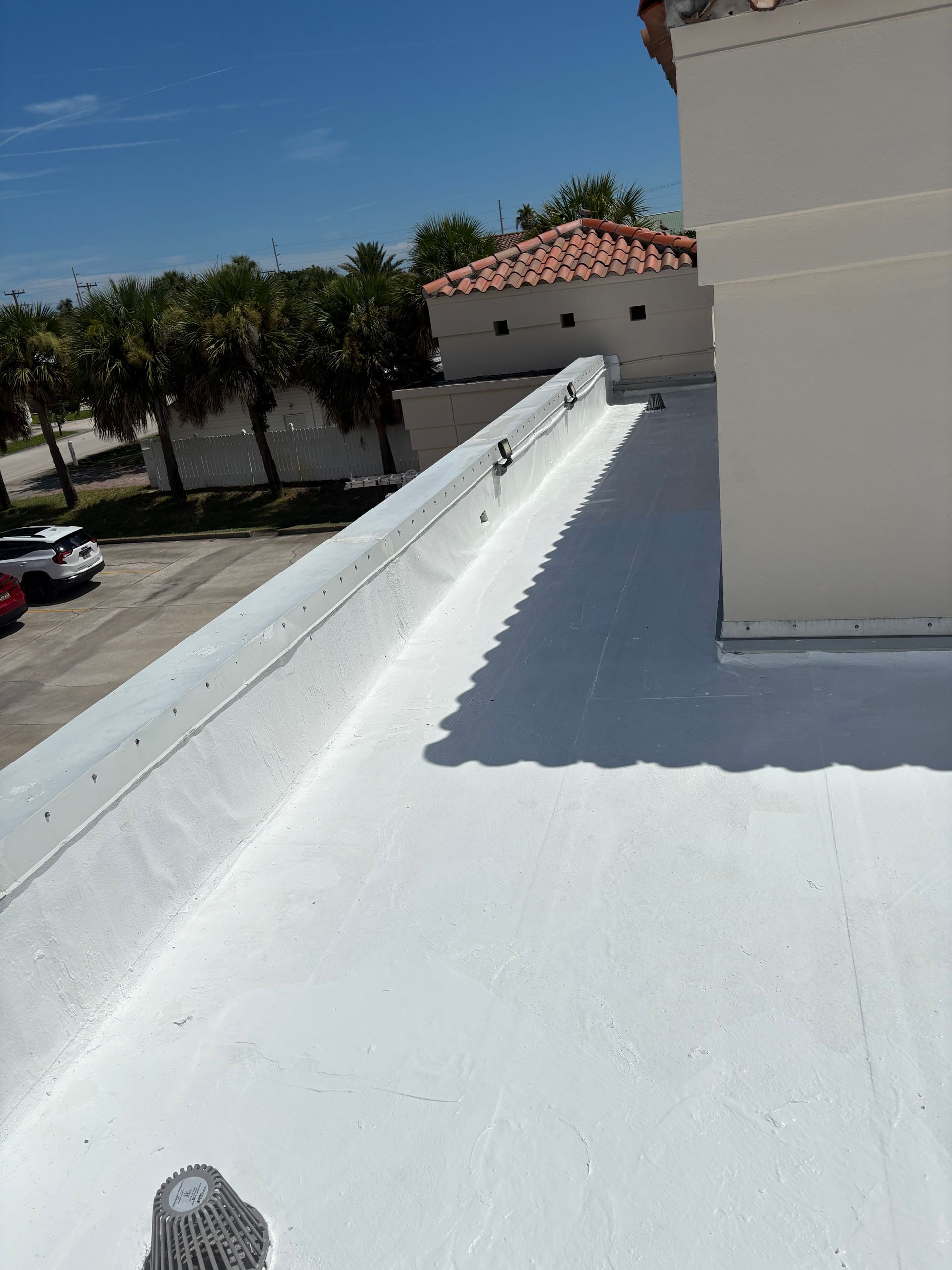 White flat roof with a parapet. Bright sunlight, clear blue sky. A parking lot and other buildings in the background.